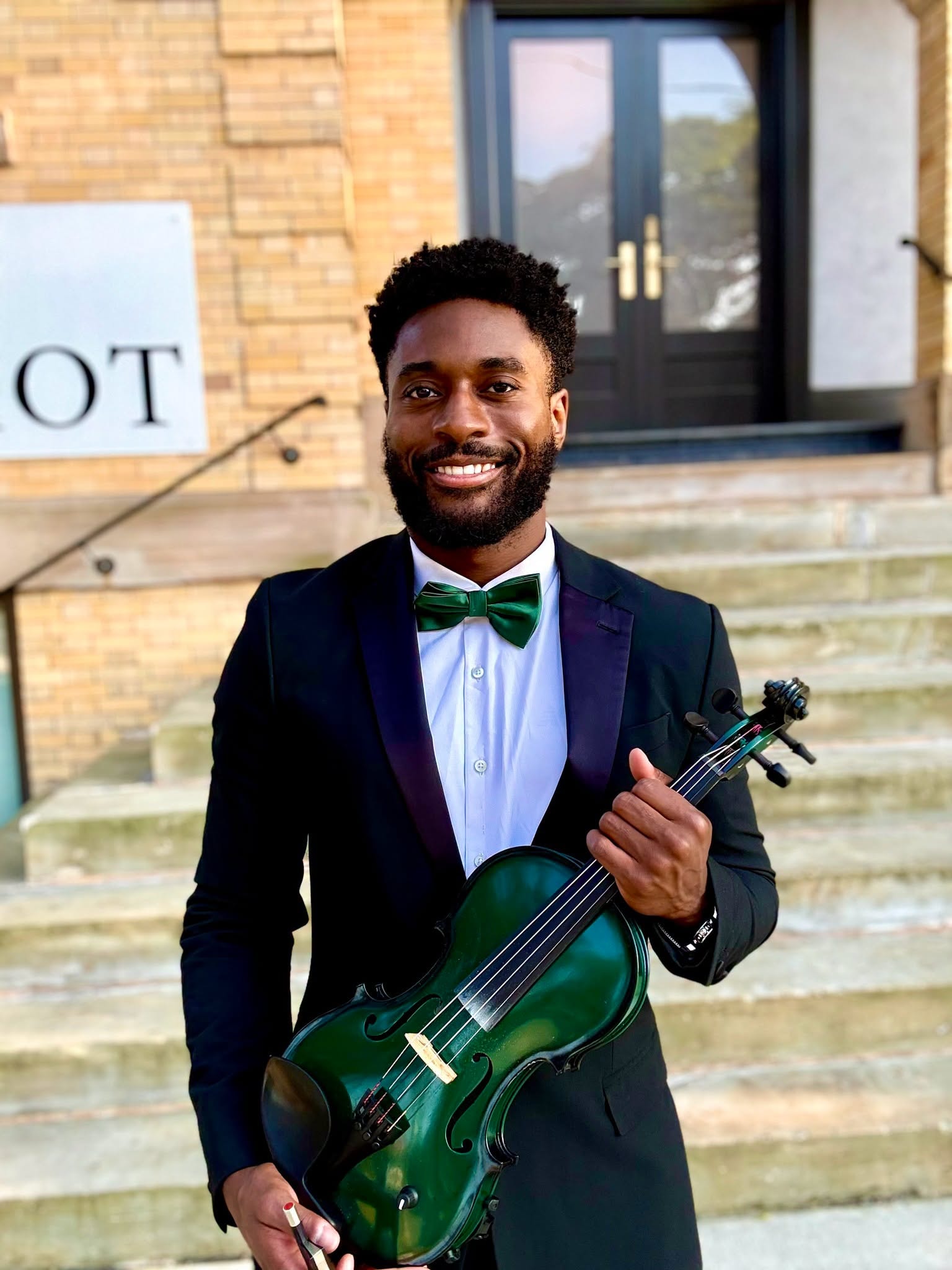Manny Apea in tuxedo holding his green electric violin on the steps outside